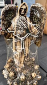 Figura esotérica de la Santa Muerte con alas de ángel extendidas, túnica beige y base decorada con calaveras y un búho.
